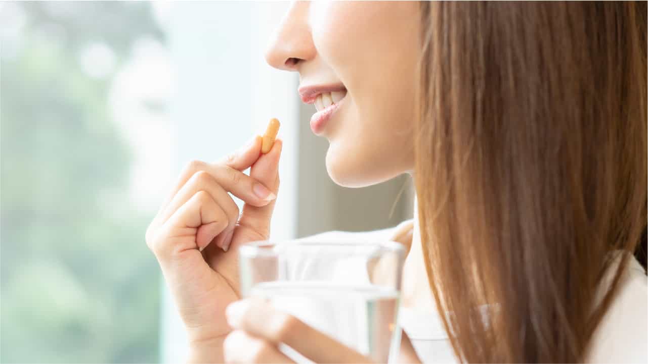 Can you take vitamin C on empty stomach? A woman holding a vitamin C capsule up to her mouth and smiling. In her other hand she holds a glass of water