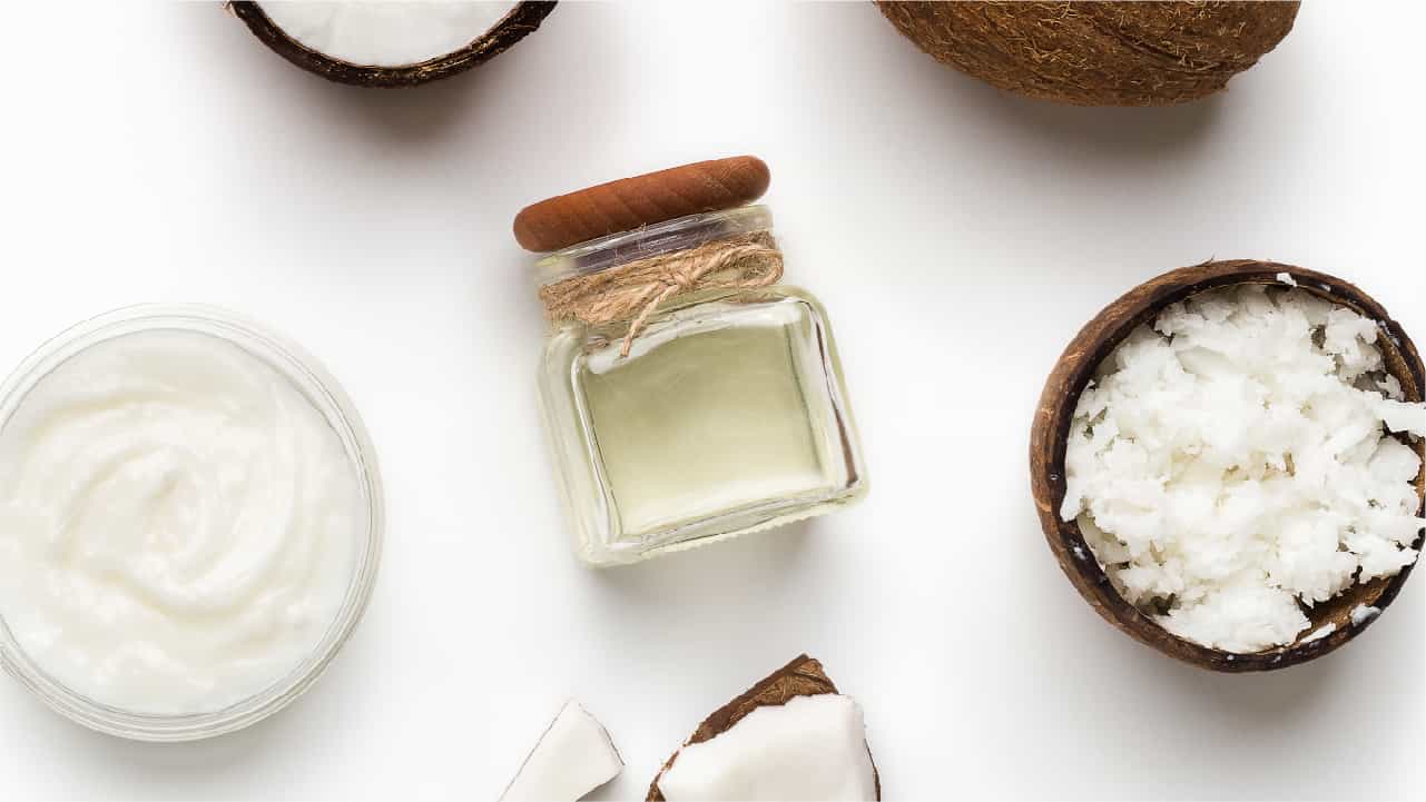 MCT Oil vs Coconut Oil: What's the Difference? A small glass bottle of pure MCT oil with a wood stopper and twine wrapped around its neck. It is surrounded by a glass bowl of coconut oil, a coconut shell filled with shredded coconut, and random pieces of cracked coconut shells and coconut flesh.
