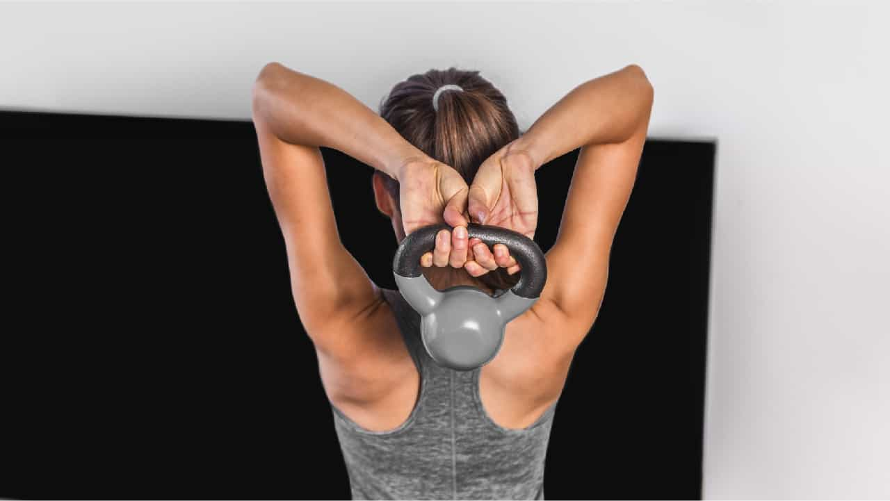 A female performing overhead triceps extension for arm toning. She holds a small kettlebell behind her head in preparation to extend it above her head.