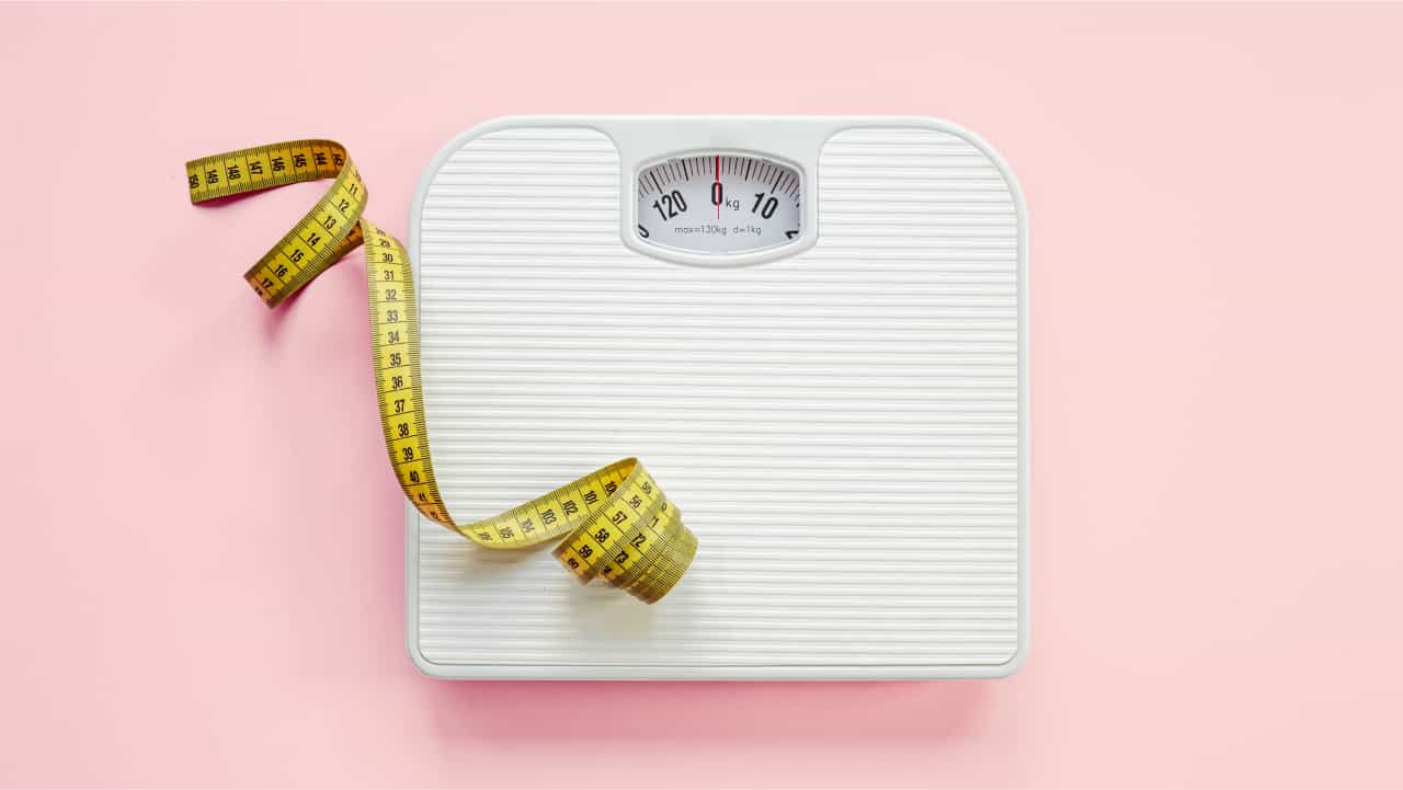 Top-rated MCT oil can help weight loss and fat burning. A white scale and yellow tape measure against a pink background.
