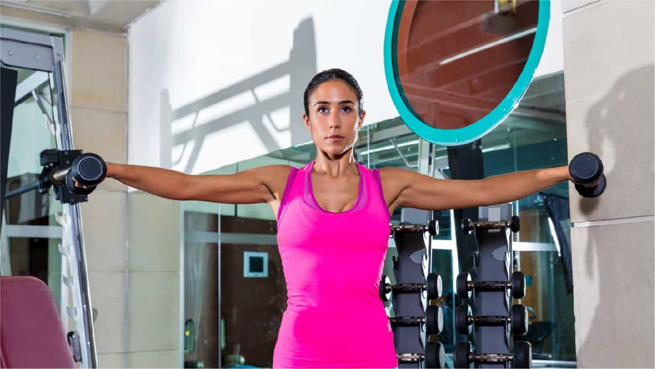 A focused woman performing lateral raises in the gym. Her well-toned arms are fully extended at her sides, a dumbbell in each of her hands