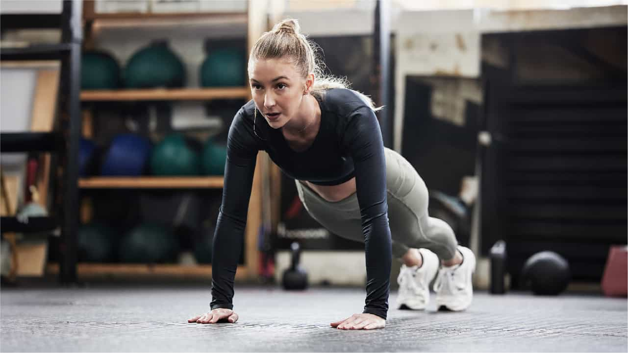 A female performing pushups in a gym. She has just pushed her body up and she has an intense look on her face