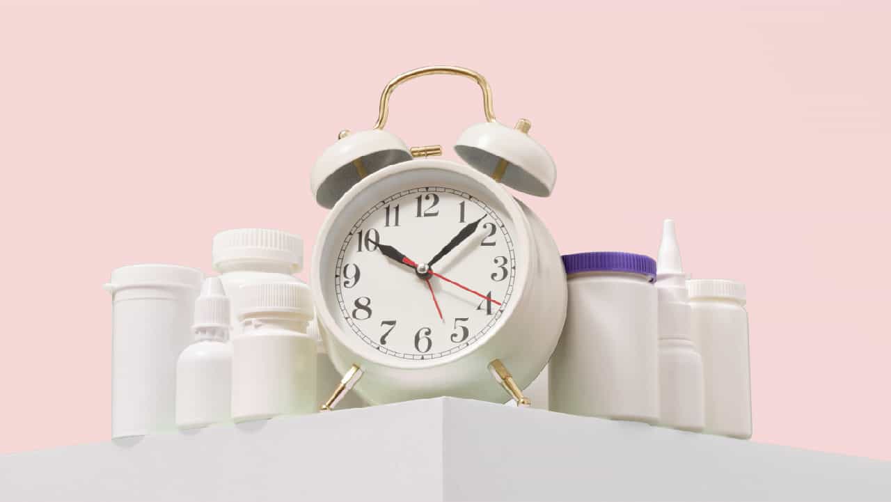 An old fashioned round faced alarm clock surrounded by supplement bottles on a pink background. Depicts asking the question how long does it take magnesium to work?