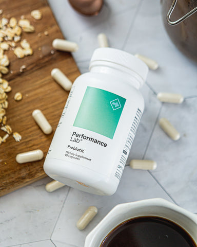 A bottle of Performance Lab Prebiotic capsules laying down on a wooden chopping board with scattered oats and a mug of coffee on a kitchen worktop.