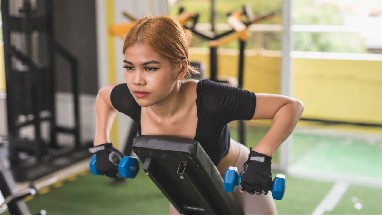 Woman with chest on an incline bench holding dumbbells and performing chest-supported row. She is in a gym with an astroturf floor.