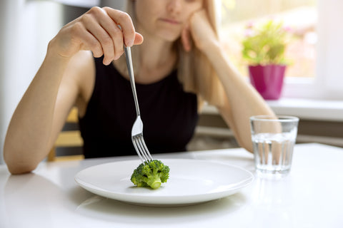 Woman looking unhappy stabbing a single piece of broccoli on an empty plate with a fork.