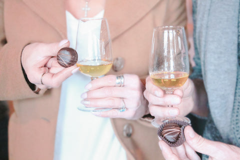 close up of a man and woman holding a glass of volcanic hills ice wine and a chocolate truffle in the other hand. Their faces are not shown. The woman is wearing a brown blazer and white shirt. The man is wearing a light blue shirt.