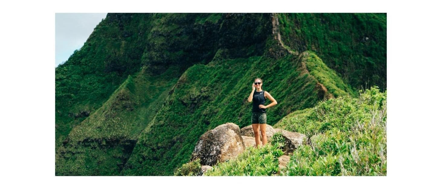 A young woman stands on a grassy mountain in Hawaii, smiling and wearing Distil Union MagLock sunglasses