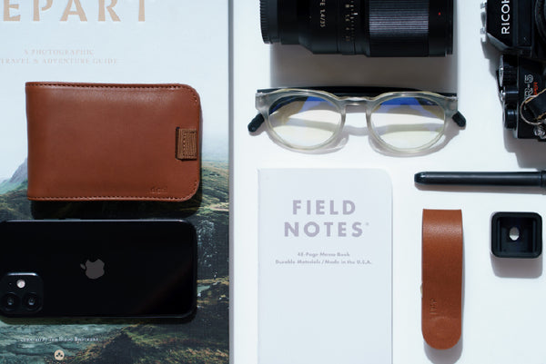 a flatlay of black, white and brown objects including a leather Wally Bifold, Field Notes, KeyLoop and Cooper ScreenSaver glasses