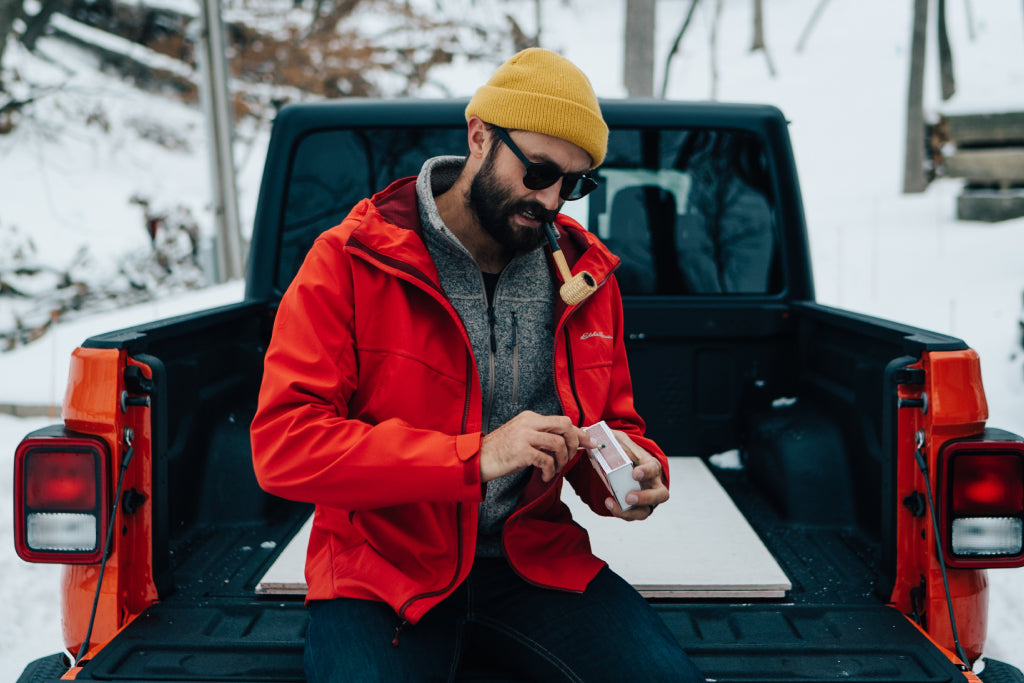 A man in a snowy place sits on the back of a truck wearing a warm hat and polarized sunglasses