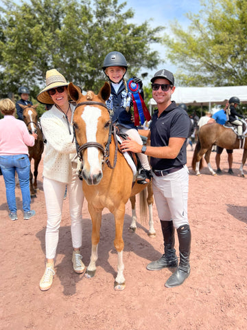 Mischa sitting on her pony, smiling with a blue ribbon. Her parents are both standing beside her smiling.