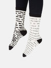 Shown on a model wearing black tights, a pair of Out of Print brand unisex mismatched cotton crew socks in white with a black heel/cuff/toe. The right sock features typewriter font listings of titles of banned books. The left sock features the same titles but blacked out.