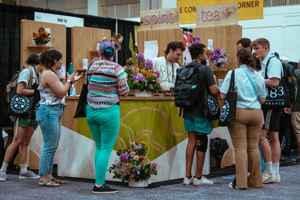 Spirit Tea's bamboo modular booth, adorned with flower arrangements by John Pendleton of Planks and Pistils. A crowd of Coffee Fest attendees gather around the booth as Taylor Cowan serves them tea. The booth presents grass green and bamboo colors. The front desk shows a white angular design with green leaf designs.
