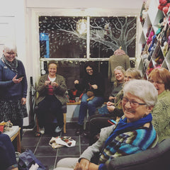 A group of women sit in All About The Yarn at night knitting and smiling