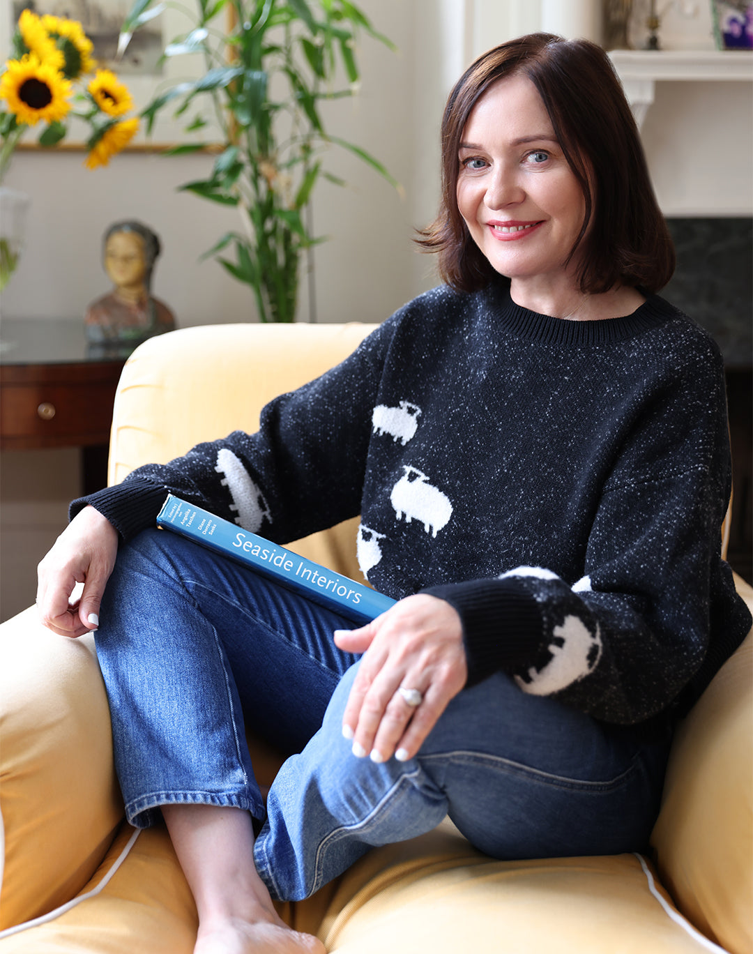 Margaret O'Leary sitting cross-legged on a yellow armchair. She is wearing a black knit jumper with white sheep on it and holding a blue book with the title "Seaside interiors" printed on the spine.