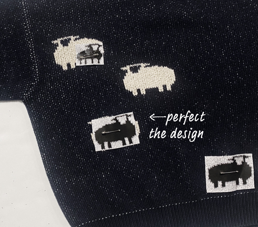 A close-up view of the sheep jumper. Small paper sheep are pinned to the sweater indicating that the placement of the sheep is being decided.