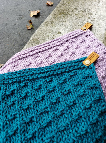 A teal and a lilac knit dishcloth lay on a cement curb.