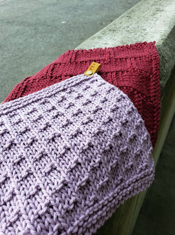 A lilac knit dishcloth lays on top of a dark red knit dishcloth on a cement curb.