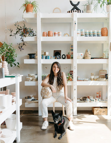 Emily Reinhardt, The Object Enthusiast, sits on a stool in her sun-lit studio in front of her studio shelves with her dog Ruby. Ruby is a medium-sized, black mixed breed dog seated on the floor in front of Emily. The shelves are filled with handmade ceramic objects made by Emily for The Object Enthusiast.