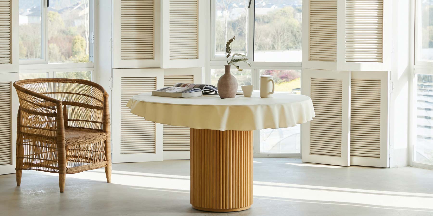 Minimalist Coastal interior. Shot frames a modern medium circular dining table. The table is covered with a short table cloth and there are two mugs, one flower vase, and one book on top of it. There is a ratan chair on the the left side of the table and the room has several open windows on the background.