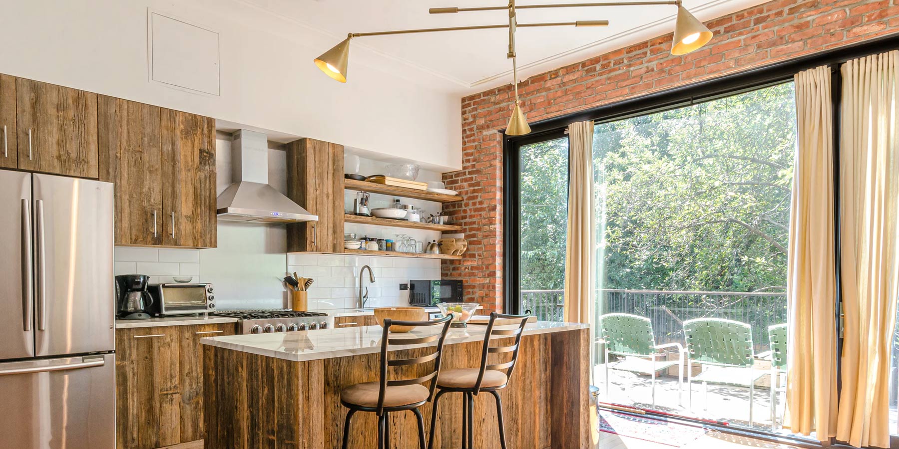 Rustic Kitchen space with reclaimed wood cabinets and kitchen island. The kitchen island has a marble top. There is a large window on the right illuminating the room. There also a modern brass (gold color) lamp on the ceiling. 