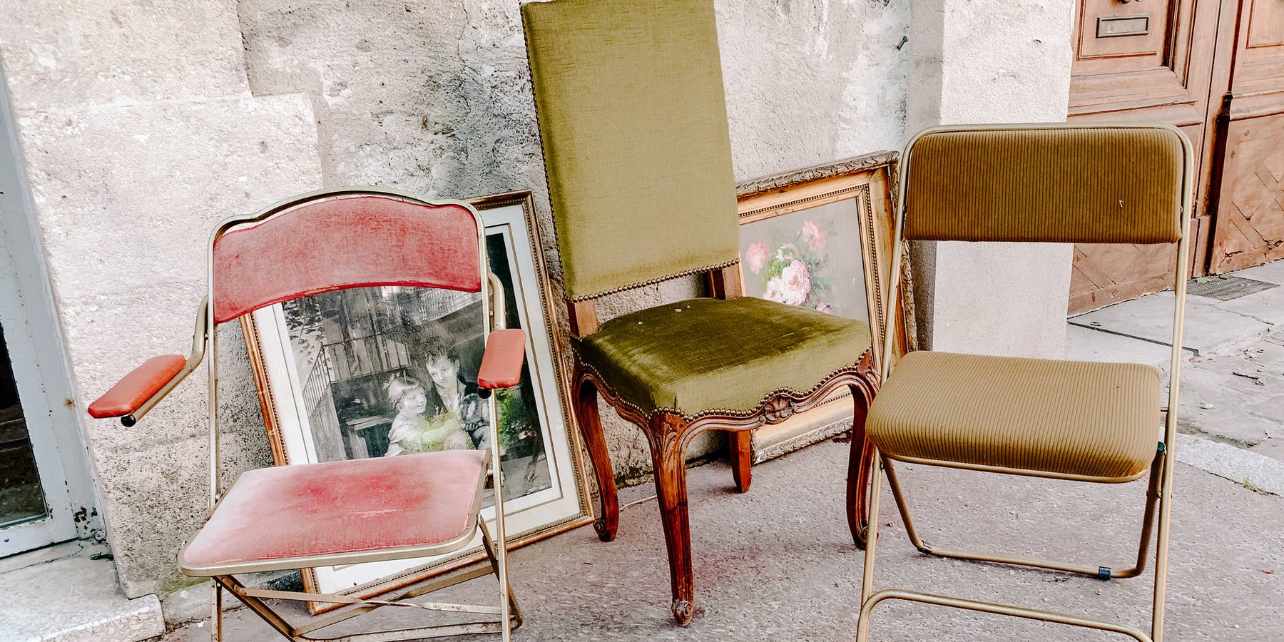 Three vintage chairs on a sidewalk. The left chair is red, the middle chair is green appostery with wooden legs and the right chair is mustard color.