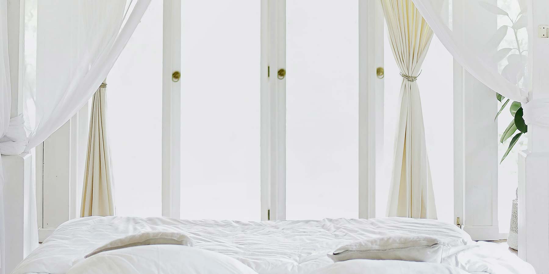 White bedroom space. The shot is facing a large window and the end or a queen size bed with white sheets. There are also 2 sets of white curtains, one for the window and one seems to be from the bed. All furniture and decoration on the shot is white, including the the walls and the curtains. 