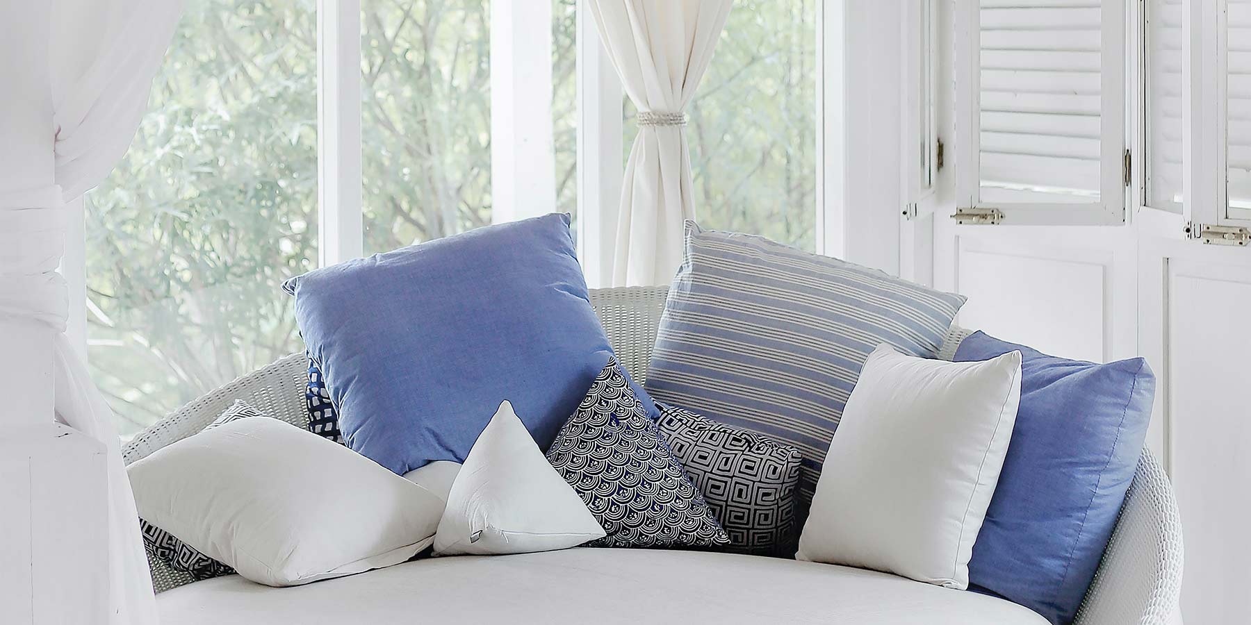 Circular beach house couch with several pillows on top of it. The pillow have different colors and patterns. The pillows very from white, light blue, and dark blue hues. The room where the couch is located is white and there is a large window or door behind it. All other elements in the background are white, including the walls, the window, and some curtains.