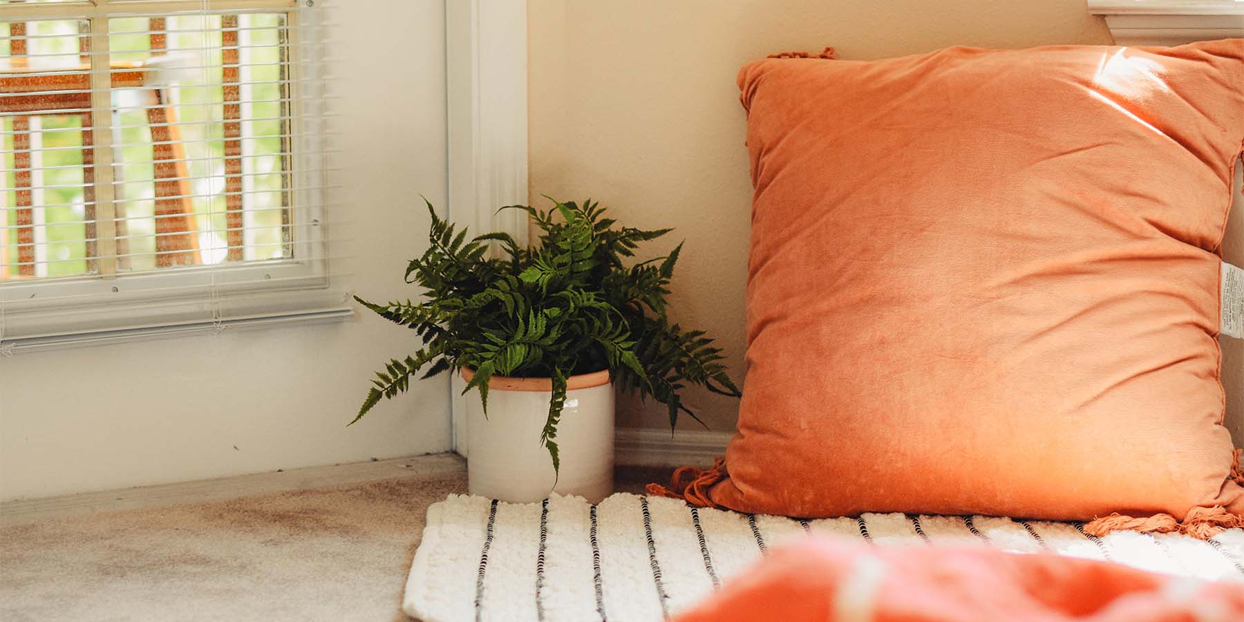 Large orange pillow on a rug on the floor next to a potted plant.