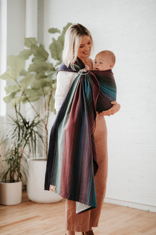 Ein Baby sitzt in einem bunten Ring Sling von Tula in Bauchtrageposition.