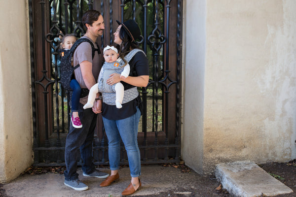 Two parents are holding hands and smiling while holding babies in tula carriers