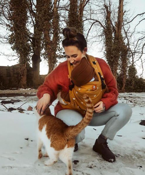 A mother carrying her child in a baby carrier while petting a cat in the snow.