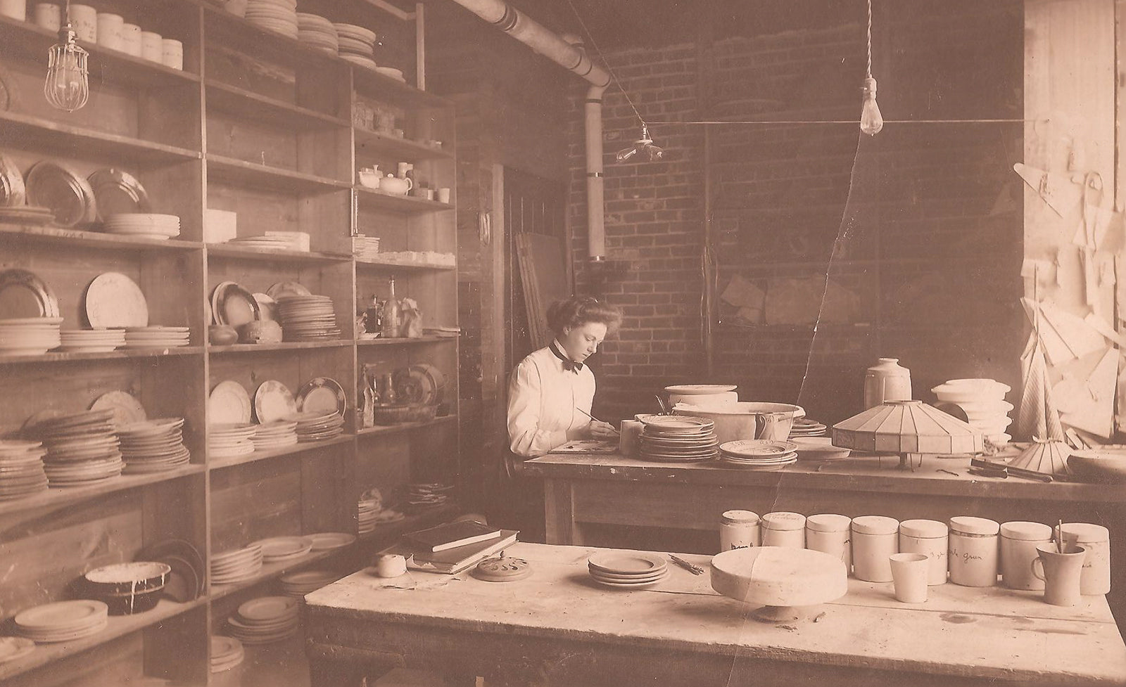 Sepia-toned photograph of Pewabic Co-Founder Mary Chase Perry Stratton working on a vessel at Pewabic Pottery in 1904. Her hair is gathered on top of her head in a neat bouffant. She wears a white, high-collared shirt with a black ribbon across the neckline.