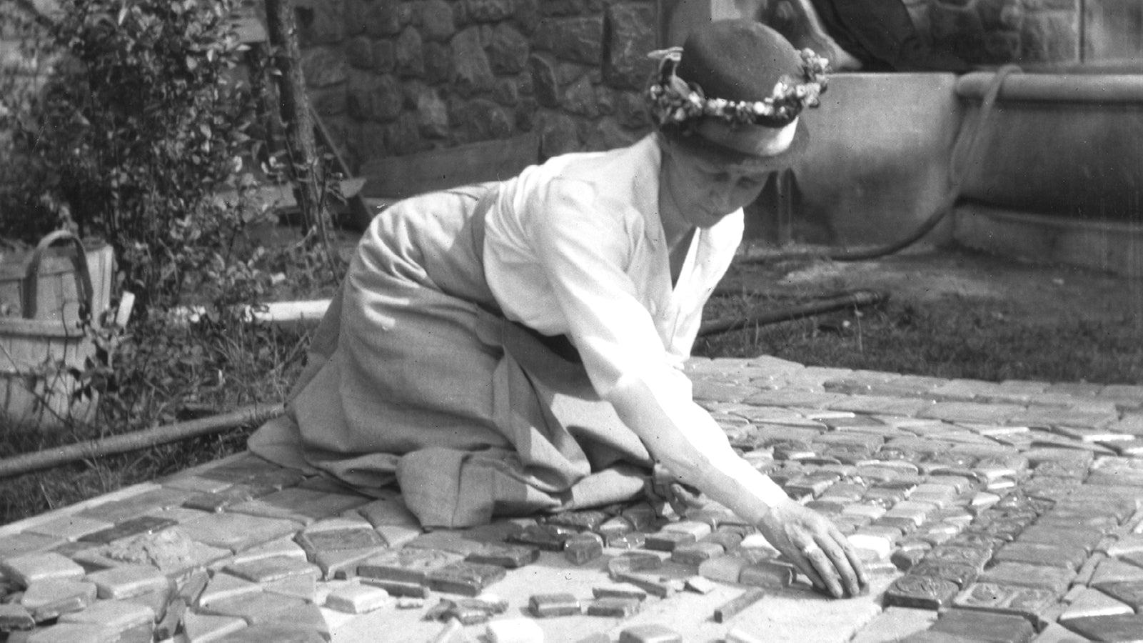 Black and white image of a woman from the 1900s in a bonnet planning a tile layout on the garden floor