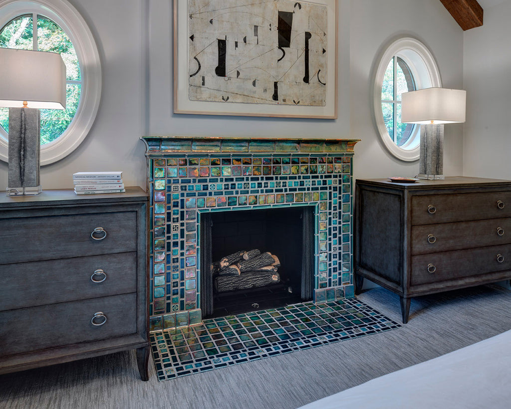 Blue and green iridescent fireplace set in a bedroom with distressed-wood dressers on either side. There are two round oculus windows above each dresser.