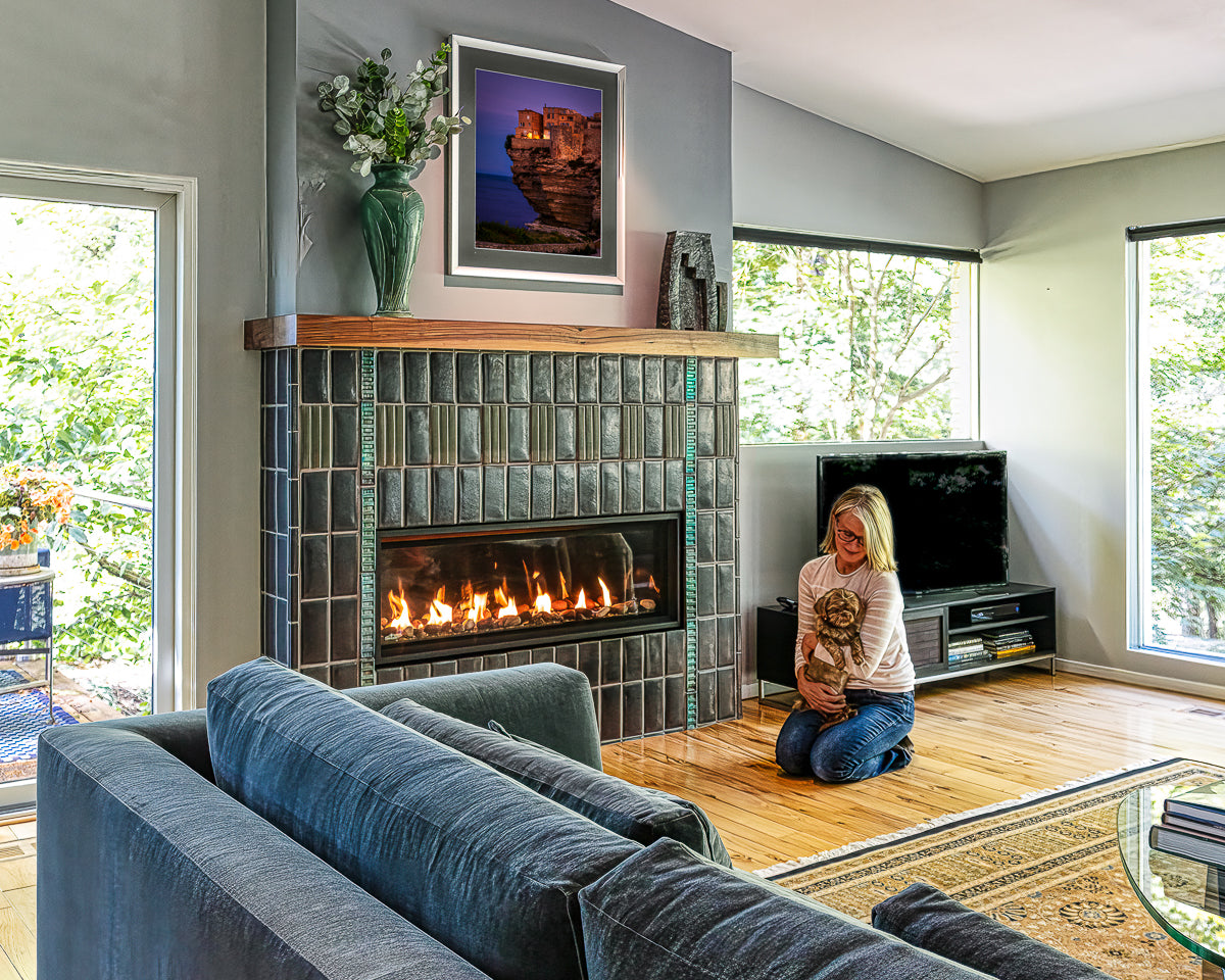 Angled view of a custom Pewabic fireplace set inside of a Midcentury-style home. There is a person holding a small dog near the lit fireplace. The windows on either side of the fireplace bring out the vibrant shine of the tilework.