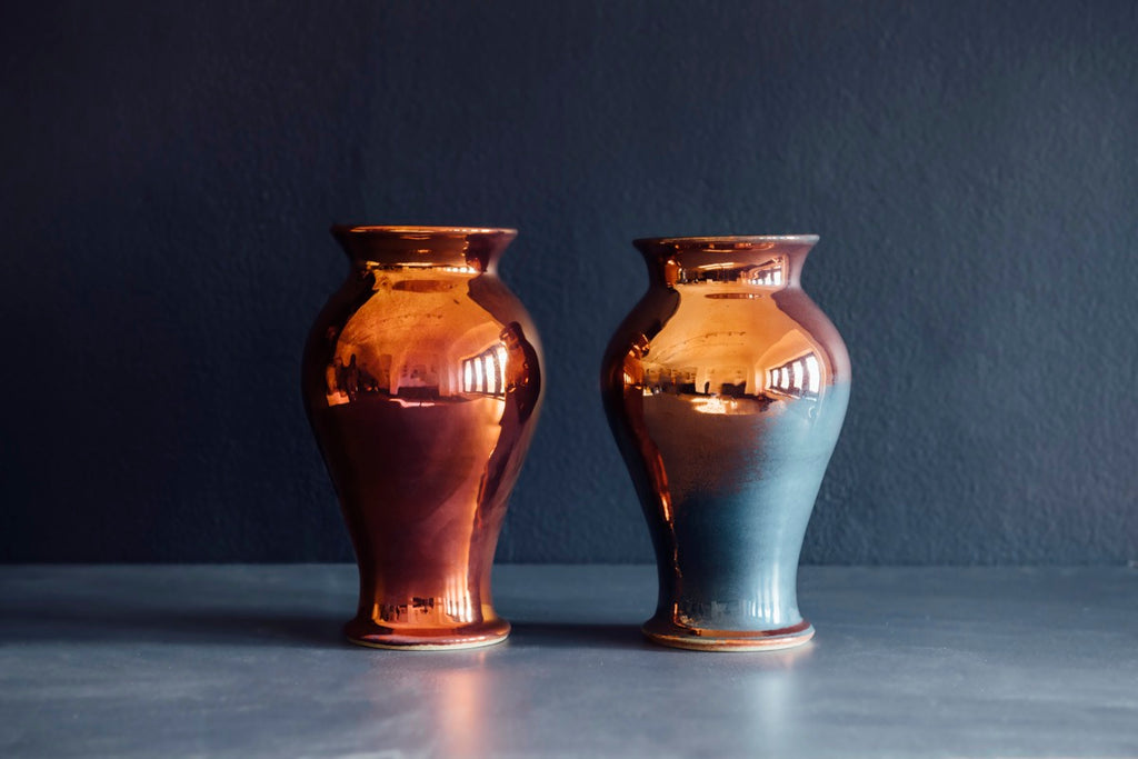 Two copper colored iridescent vases side by side against a dark gray/blue background. The vase on the left looks like a vibrant sunset, while the vase on the right has flashes of muted blue on the surface of a mostly copper base.