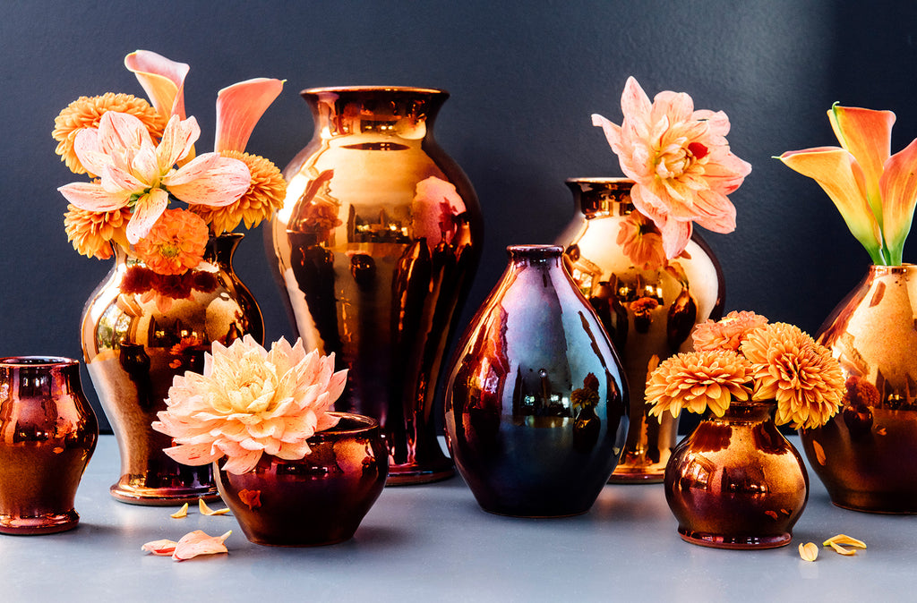 Various copper iridescent vases lined up against a dark blue backdrop. The vases are shown in a range of sizes. Some of them are holding pale pink and peach colored flowers.