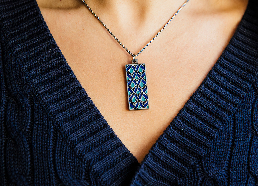 The Women's City Club necklace is a rectangular metal pendant with a geometric design that includes teardrop shapes and smoothly curved lines in various shades of blue. The pendant hangs from a silver chain around the neck of a woman in a navy cable knit sweater.