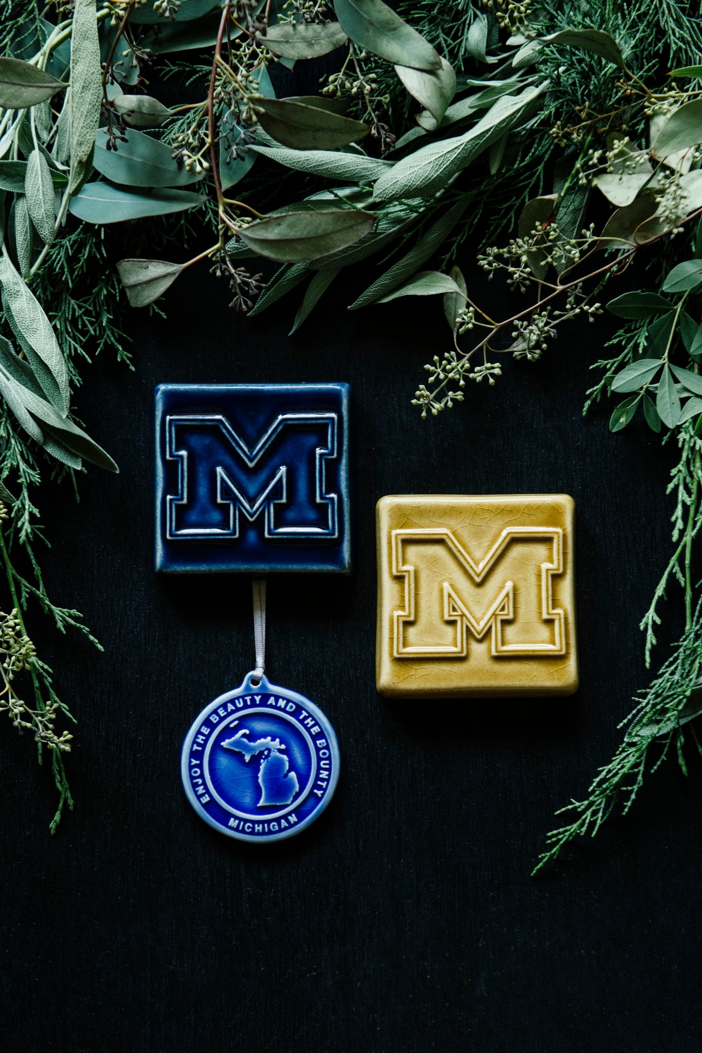 Two 4x4 tiles in a dark, glossy blue glaze and bright, glossy yellow glaze showcasing the U-M logo. There is also a true-blue ornament with an outline of the state of Michigan that reads "The Beauty and the Bounty".