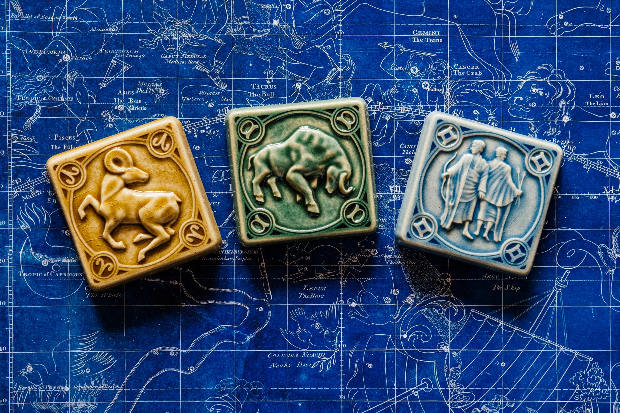Our hand-sculpted Aries, Taurus, and Gemini tiles are placed in an arch on a deep, blue, historic star map. The Taurus tile features a sturdy bull with its head bowed and horns lowered. The Aries tile has an alert ram with its legs positioned as if it is prancing. The ram's head is turned backwards over its shoulder. Gemini is depicted by two men, “the twins” in flowing robes. One is holding a bow, the other is holding two arrows.