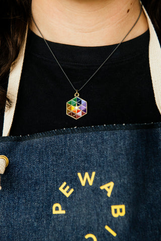 Close up photo of a Pewabic employee wearing a denim apron with a bright, yellow Pewabic logo. They are wearing a colorful hexagonal necklace with bronze detailing.