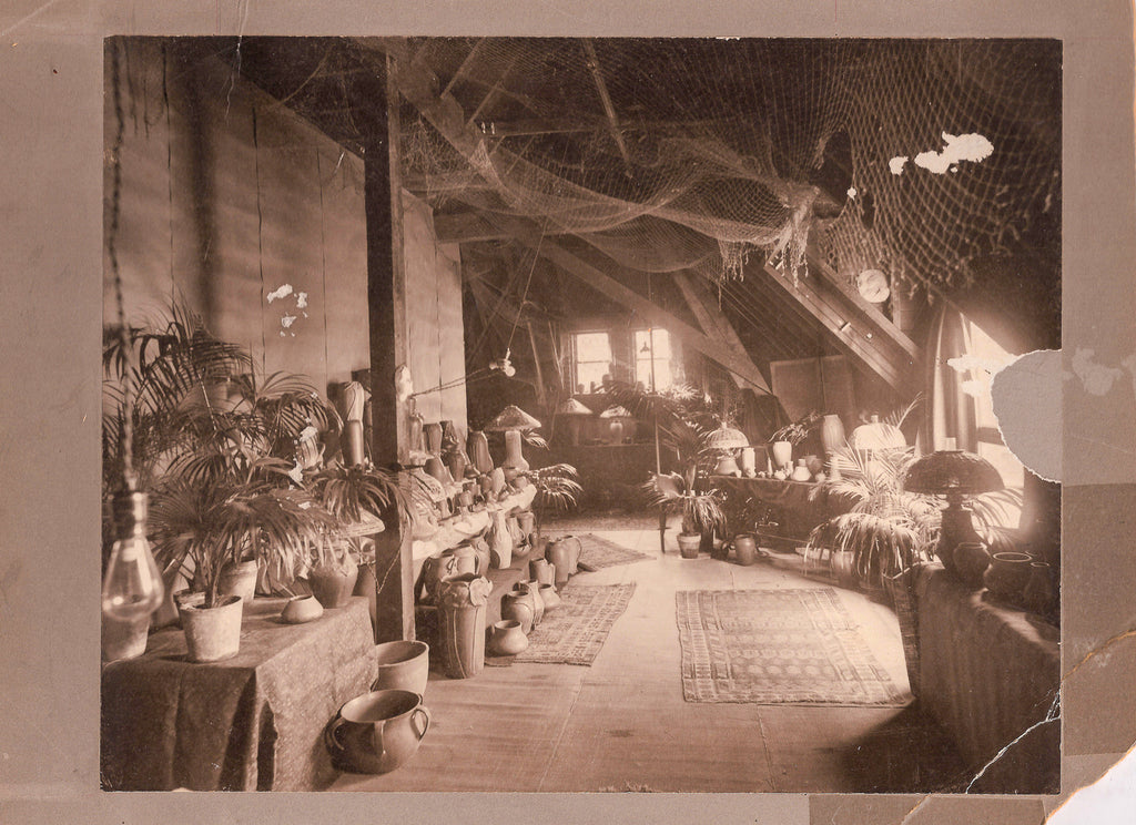 Dark sepia film photo scan of a exhibition that took place in the Stable Studio in 1903. There are ceramic pieces lining the left wall alongside tropical-looking plants and foliage.