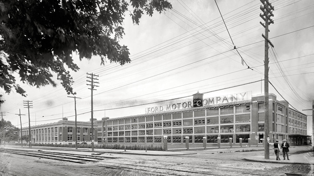 Exterior shot of the Ford Motor Company’s Highland Park Plant, birthplace of the Ford Model T in the year 1910. Two workers wearing pork pie hats and suits stand adjacent to the building near railroad tracks.