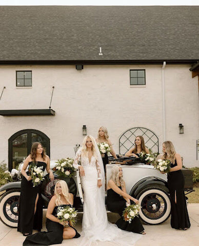 Blonde bride in a lacy, long-sleeved white gown posing in front of a vintage car with her six bridesmaids dressed in black.