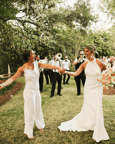 Two beautiful brides in white holding hands, one wearing a dress, one wearing a pantsuit.