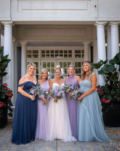 Bride in white dress posing between two bridesmaids in lavender dresses, one in a navy blue dress, and one in a light blue dress