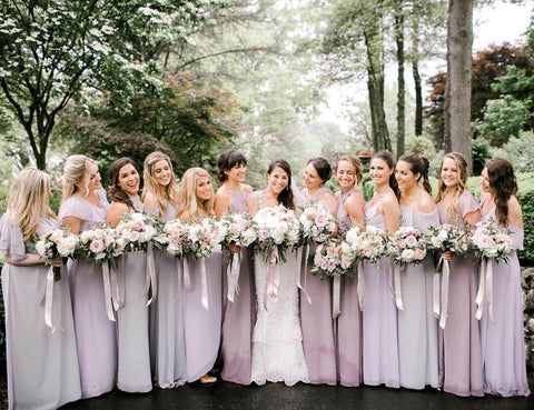 Bride in white dress with six bridesmaids in lavender dresses on each side of her. All of them are holding white and pink bouquets with lavender ribbons.