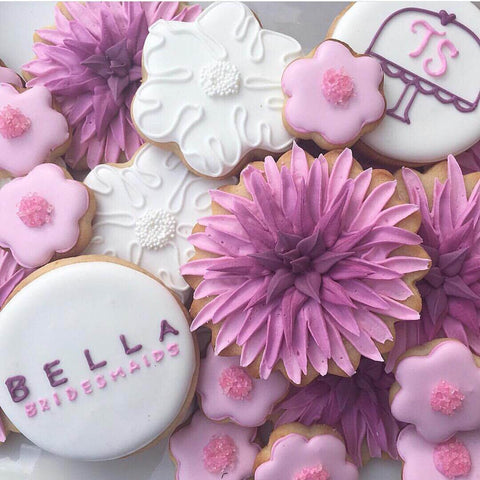 A spread of white, pink, and lavender-frosted cookies. One of the cookies says, "Bella Bridesmaids" in purple and pink frosting on a white cookie.