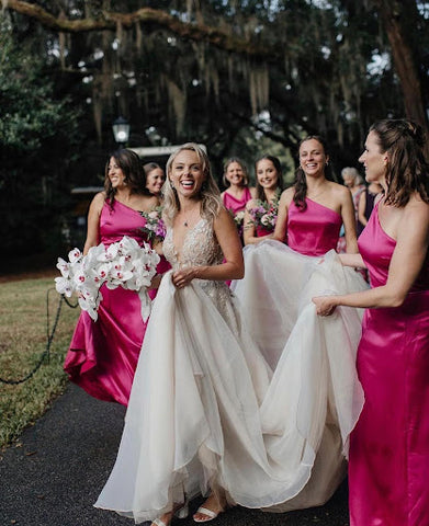 Smiling bride walking while a few of her bridesmaids (in fuschia gowns) carry her train.